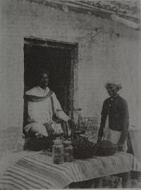 A shopkeeper seated in a doorway behind a counter with a hand balance, glass jars and bowls of dark loose material, a second man standing beside it.