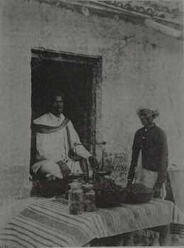 Two men at a whitewashed shopfront, one seated in the doorway beside a hand balance, with stoppered glass jars and two shallow metal bowls of dark loose material set out on a striped cloth over the counter.