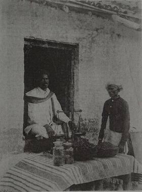 Two men at a whitewashed shopfront, one seated in the doorway beside a hand balance, with stoppered glass jars and shallow metal bowls of dark loose material on a striped cloth.