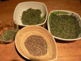 Four bowls on a wooden table, one holding brown cannabis seeds and three holding dried plant material.