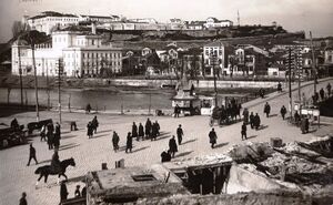 A paved square by a river with people on foot, a horse-drawn carriage and a rider, a bridge on the right and fortress walls on the hill above