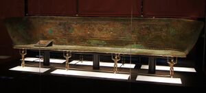 A long bronze couch with a high back, raised on small cast figures, in a dark museum case