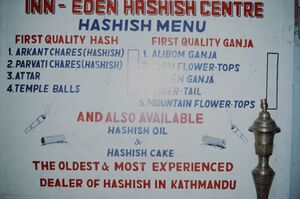 A painted menu board headed Inn-Eden Hashish Centre listing four kinds of hashish and five of ganja, with hashish oil and hashish cake below, and a brass stand in front
