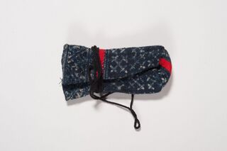 A small folded pouch of dark blue cloth patterned with pale crosses and spots, with two strips of red cloth, tied round with a black cord whose loose end hangs in a loop